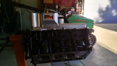 1985 Jeep CJ7 - A black V8 engine block is mounted on an orange engine stand, with a silver oil pan and a portion of a green lawn tractor visible in the ba…