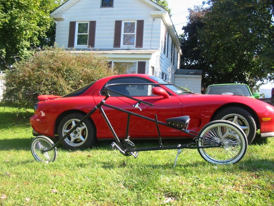 Custom black stretched lowrider bicycle on grass in front of a red sports coupe and white house