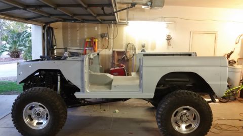 1979 Ford Bronco - A light gray, unfinished Ford Bronco body sits in a garage, lifted high on large off-road tires. The vehicle is missing its doors and roof,…