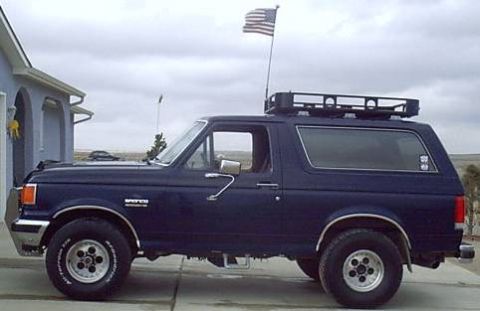 1988 Ford Bronco - Dark blue 1988 Ford Bronco parked outside a house, equipped with a roof rack and an American flag.
