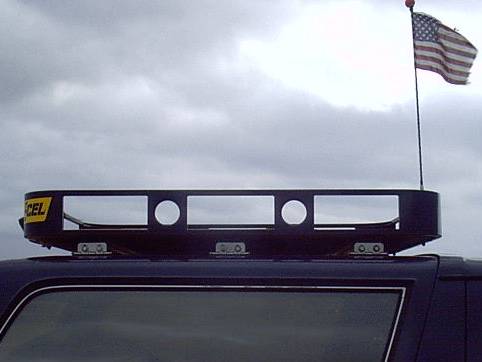 Black roof rack mounted above a vehicle roof with round light cutouts and a yellow sticker, American flag and cloudy sky behind