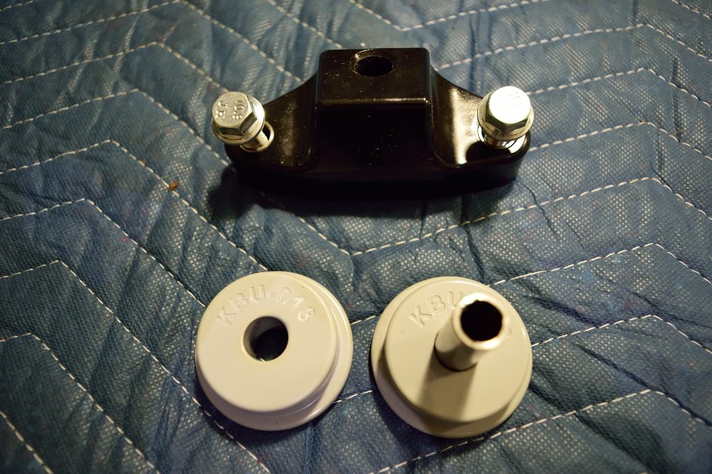 Black metal bracket with bolts and two gray bushings labeled KBU-018