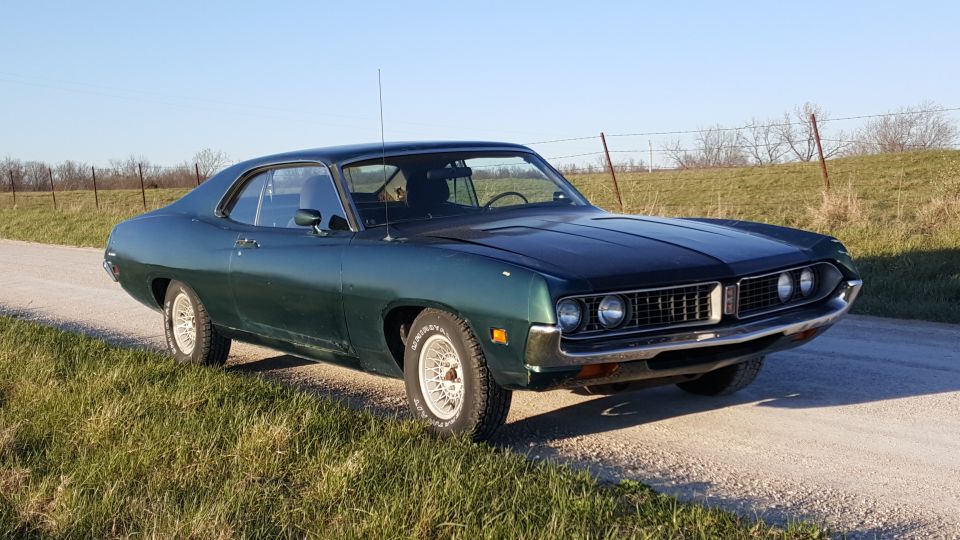 1971 Ford Torino coupe, dark green, parked on gravel road with grassy field.