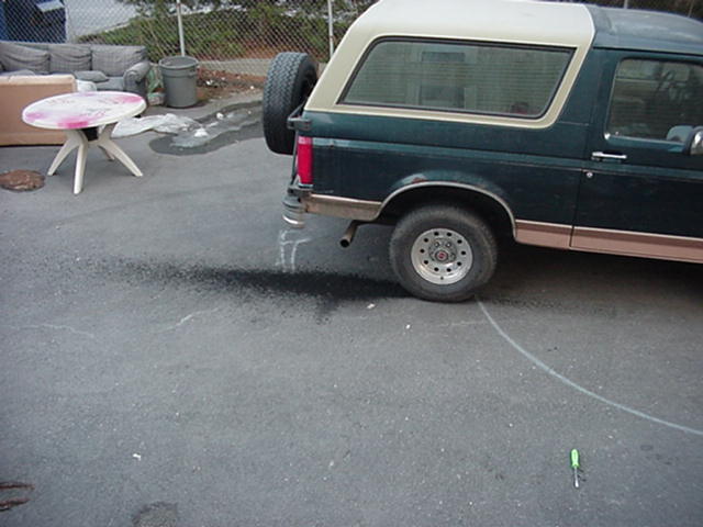 Rear quarter of a green Ford Bronco with tan camper top and swing-out spare tire in a paved yard with discarded furniture