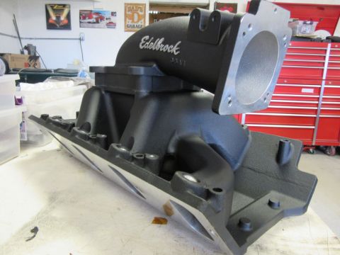 Black Edelbrock intake manifold with silver flange on a table in a workshop.