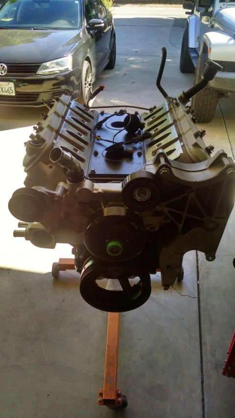 1985 Jeep CJ7 - A V8 engine is mounted on an orange engine stand, with a black Volkswagen Jetta and a silver Jeep visible in the background.