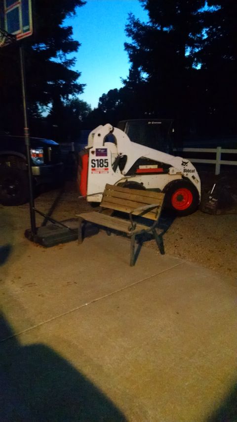 1985 Jeep CJ7 - A white Bobcat S185 skid steer loader is parked on a concrete driveway next to a wooden bench and a basketball hoop. The sky is a deep blue…