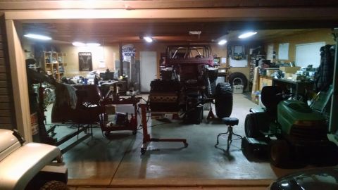 1985 Jeep CJ7 - A garage scene shows a partially disassembled Jeep CJ7 with its engine out, sitting on an engine stand. A green riding lawn mower and vario…