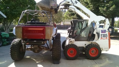 1985 Jeep CJ7 - A heavily modified red Jeep CJ7 with a roll cage is positioned next to a white Bobcat S185 skid steer loader. The skid steer's bucket is ra…