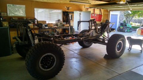 1985 Jeep CJ7 - A custom-built off-road vehicle chassis with large tires is parked inside a garage. A black dog stands to the right of the vehicle.