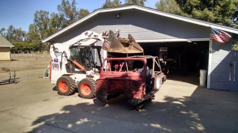 1985 Jeep CJ7 - A red Jeep CJ-style body is being lifted by a Bobcat skid steer loader with a large bucket attachment. The Jeep body is partially disassemb…