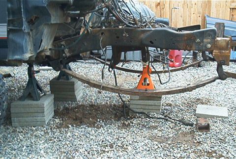 1986 Ford Bronco - Underside view of a vehicle frame supported on stacked concrete pavers and an orange jack stand, with a curved leaf spring visible and vari…