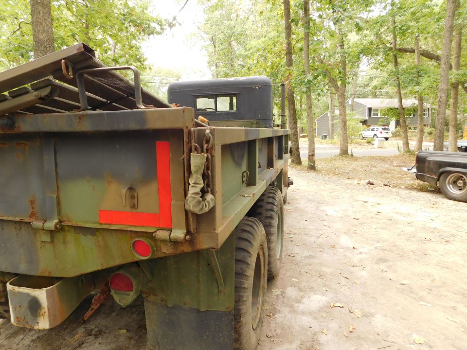 Rear side view of a weathered olive drab military truck with camouflage paint and large tires.