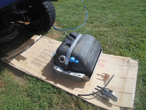 1972 Ford Bronco - A black plastic fuel tank is shown resting on a piece of cardboard on the grass. A metal strap secures the tank, and a blue hose is attache…