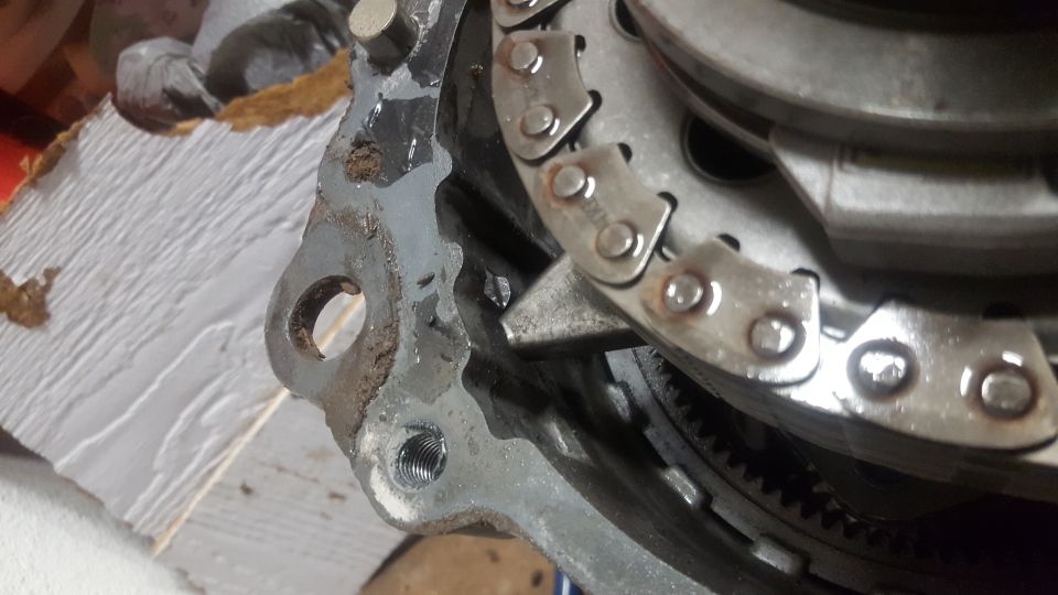 Close-up of a greasy clutch pressure plate assembly with riveted straps and threaded hole, on a wood board