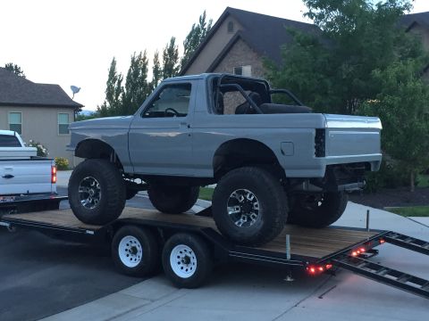 1988 Ford Bronco - A lifted, two-door Ford Bronco in primer grey is loaded onto a flatbed trailer. The Bronco has large off-road tires and a roll cage visible…