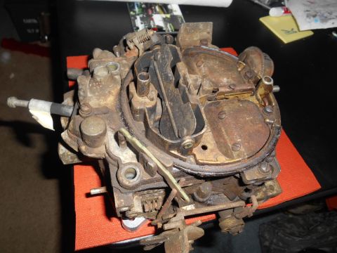1972 Ford Bronco - A rusty, old carburetor sits on an orange mat on a black surface. The carburetor has multiple metal components, springs, and screws visible.