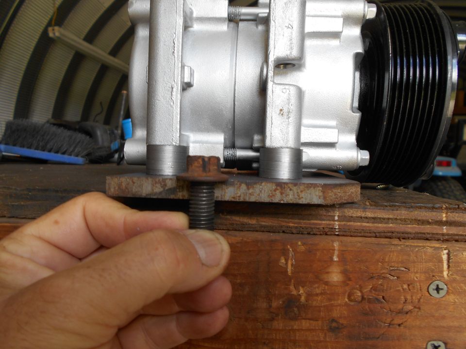 Hand holding bolt in front of silver AC compressor with black pulley.