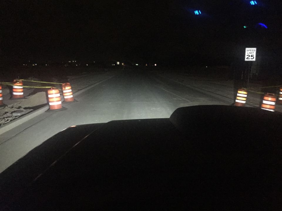 Dark car hood with headlights illuminating a road with construction barrels and a speed limit sign