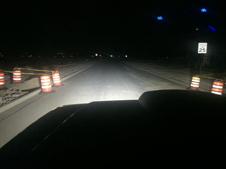 Vehicle hood at night with headlights illuminating a road with construction barrels