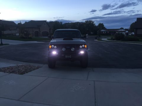 1988 Ford Bronco - A dark Ford Bronco is parked in a driveway at dusk, its headlights and auxiliary lights illuminated.