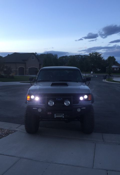 1988 Ford Bronco - This lifted SUV features an aftermarket bumper, winch, and auxiliary lights, with its headlights illuminated.