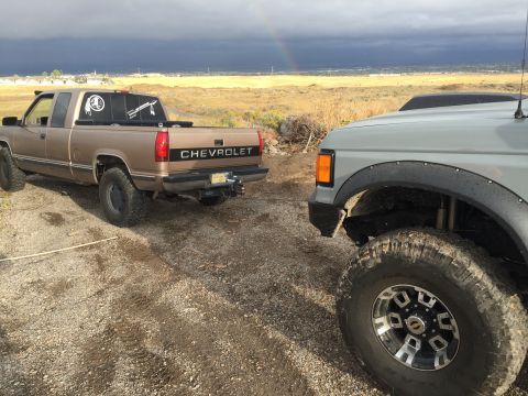 1988 Ford Bronco - Two trucks are parked on a gravel road under a stormy sky with a rainbow visible.