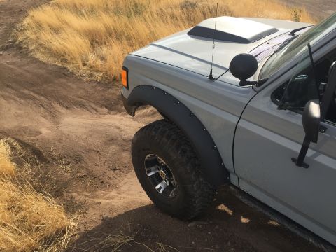 1988 Ford Bronco - A gray SUV with fender flares and a hood scoop drives on a dirt road.