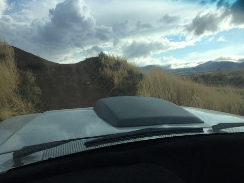 1988 Ford Bronco - Vehicle with a hood scoop navigates a dirt road through a landscape of dry grass and hills under a cloudy sky.
