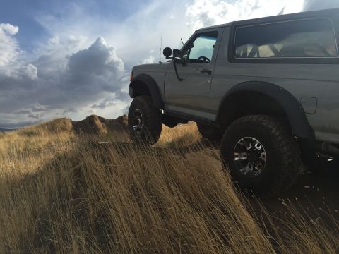 1988 Ford Bronco - This Ford Bronco is captured mid-climb on a dry, grassy slope with dramatic clouds overhead.