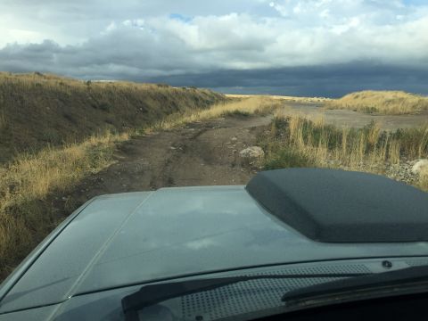 1988 Ford Bronco - View from inside a vehicle with a prominent hood scoop, driving on a rough dirt track under a cloudy sky.