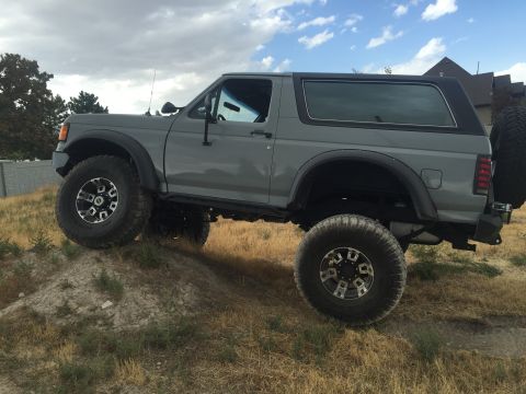 1988 Ford Bronco - A gray Ford Bronco is shown from a three-quarter front angle, positioned on a dirt mound with a cloudy sky above.