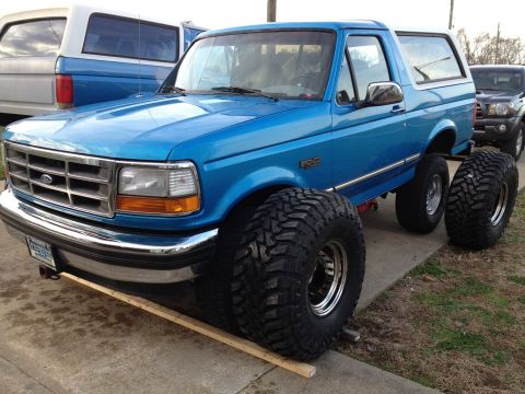 1995 Ford Bronco - A lifted blue Ford Bronco with oversized tires is parked on wooden ramps, with another vehicle visible behind it.
