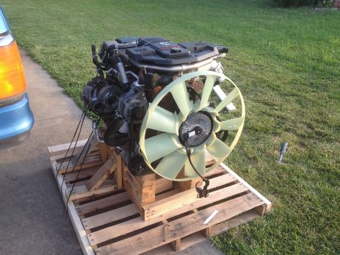 1995 Ford Bronco - An engine is mounted on a pallet, with a large fan attached, beside a blue vehicle's front light.