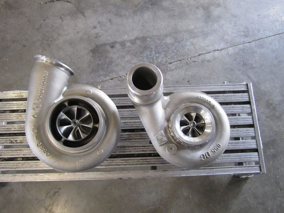 Two silver turbochargers with visible fan blades sit on a metal grate. One turbocharger is larger than the other