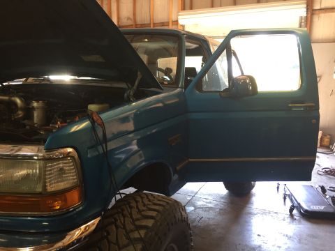 1995 Ford Bronco - This blue Ford truck has its hood open and driver's door ajar, revealing engine details and large tires.