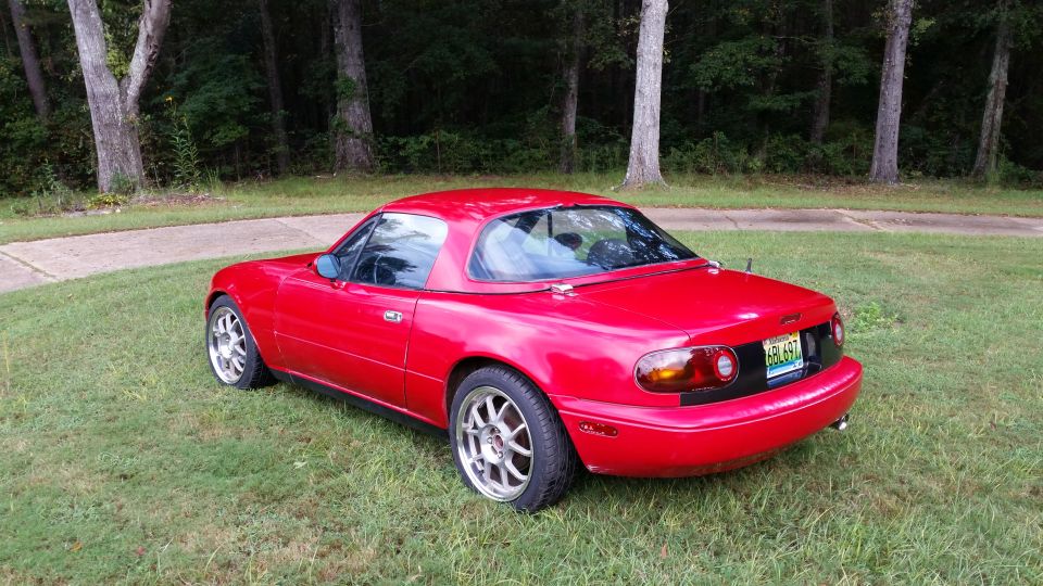 Red Mazda Miata with hardtop parked on grass, rear driver's side view.
