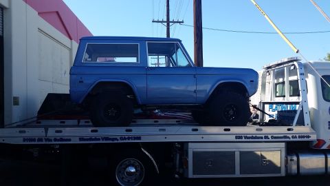 1973 Ford Bronco - A blue 1973 Ford Bronco is loaded onto a flatbed tow truck. The Bronco is seen from the side, with its tires and lower body visible.