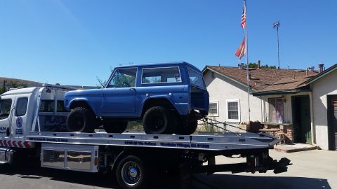 1973 Ford Bronco - A blue, two-door Ford Bronco is being transported on a flatbed tow truck. The Bronco is positioned on the rear of the truck, with its large…