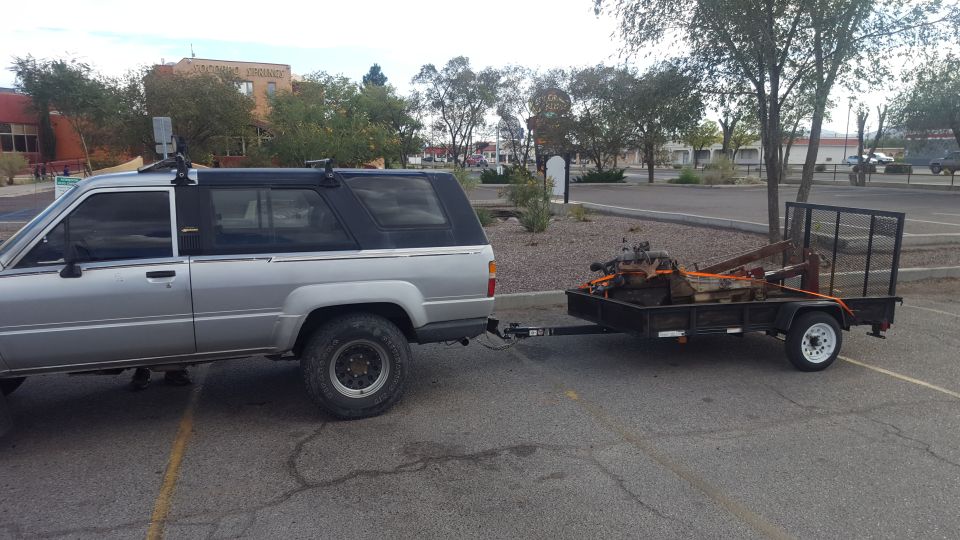 Silver compact SUV with black top towing a black utility trailer loaded with an engine in a parking lot