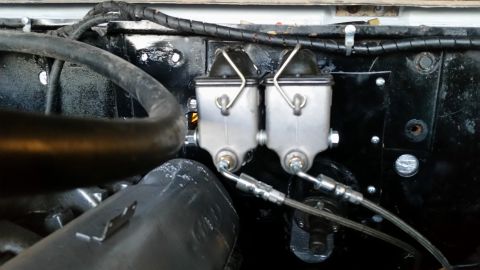 1979 Ford Bronco - A close-up view of a dual brake master cylinder assembly mounted on a black firewall, with brake lines and hoses visible.