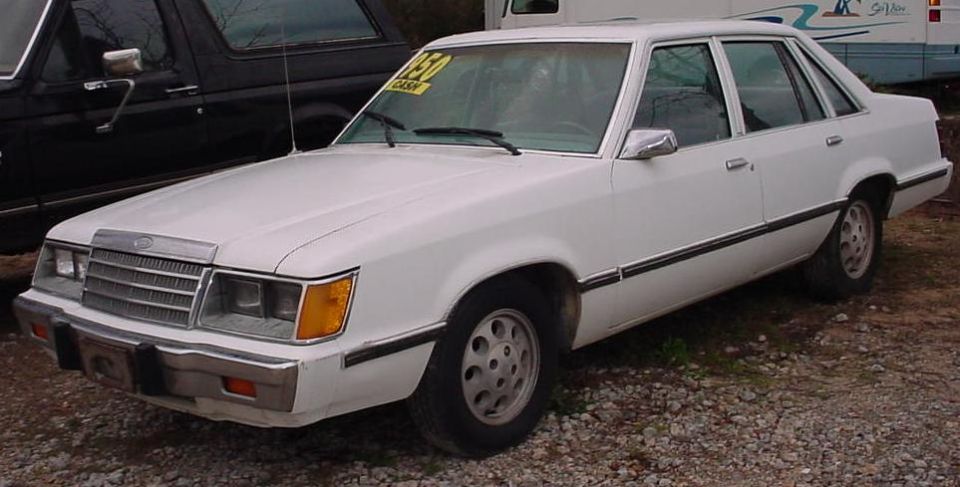 White 1984 Ford LTD sedan, front 3/4 view on gravel lot with yellow $950 cash for-sale sign in windshield