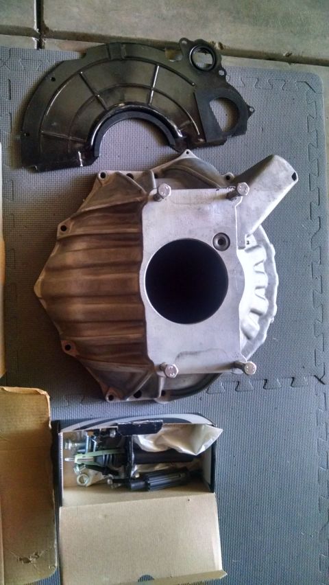 1985 Jeep CJ7 - A transmission bell housing and a black metal dust shield are laid out on a grey mat. Below them, a cardboard box contains various metal pa…