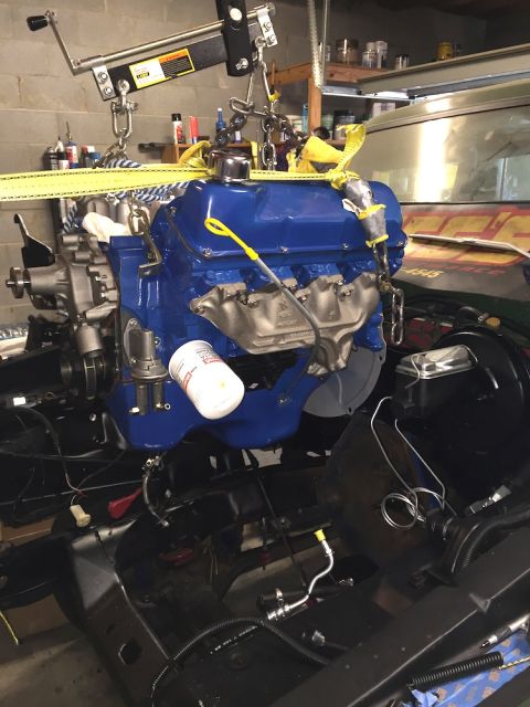 1978 Ford Bronco - A blue Ford V8 engine is suspended by a hoist and straps over a black chassis. A white oil filter is attached to the side of the engine.