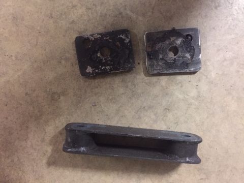 1982 Ford Bronco - Three metal parts are laid out on a concrete floor. Two square metal plates with holes are at the top, and a U-shaped metal bracket is at t…