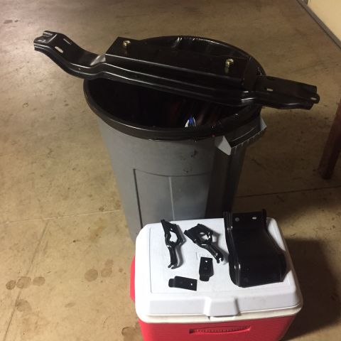 1982 Ford Bronco - Several black metal automotive parts are laid out on a red and white cooler, with a larger black metal part resting on top of a gray trash…