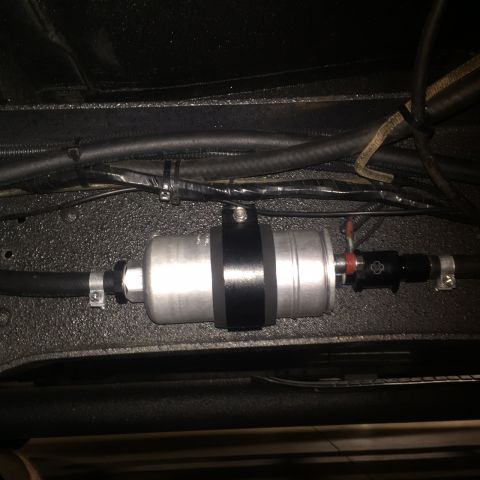 1982 Ford Bronco - A close-up view of a silver fuel filter with black bands, mounted with hoses and clamps to the underside of a vehicle.