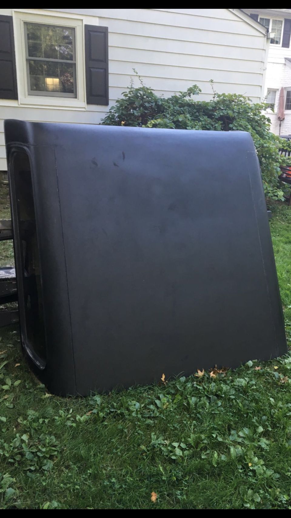 Black truck cap with a window opening, resting on grass in front of a house.