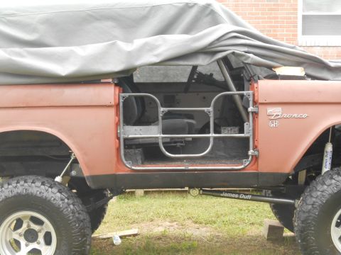 1972 Ford Bronco - The side view of a classic Ford Bronco with its doors removed and replaced with tubular roll cage doors. A grey tarp is draped over the veh…