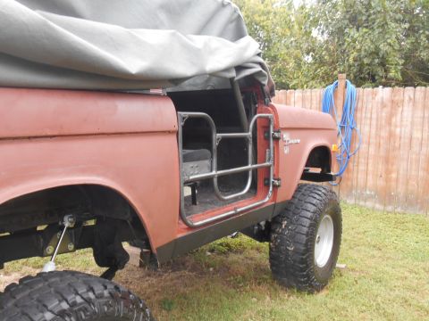 1972 Ford Bronco - The side view of a red 1972 Ford Bronco with a grey tarp covering the top. The vehicle has large off-road tires and exposed suspension comp…
